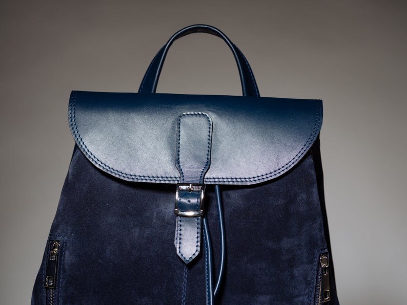 May include: A dark blue suede backpack with a leather flap and buckle closure. The backpack has a top handle and zippered pockets on the sides.