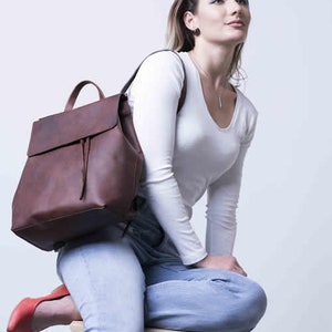 以下が含まれることがあります： 青いジーンズと白い長袖シャツを着た女性が木製の椅子に座っています。彼女はフラップとドローストリングクロージャーが付いた茶色の革のバックパックを背負っています。