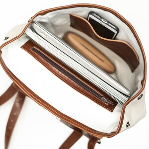 May include: The inside of a brown leather and white canvas backpack. The backpack has a zippered pocket and a large compartment that can fit a laptop and other items. The backpack also has a small pocket for a phone.