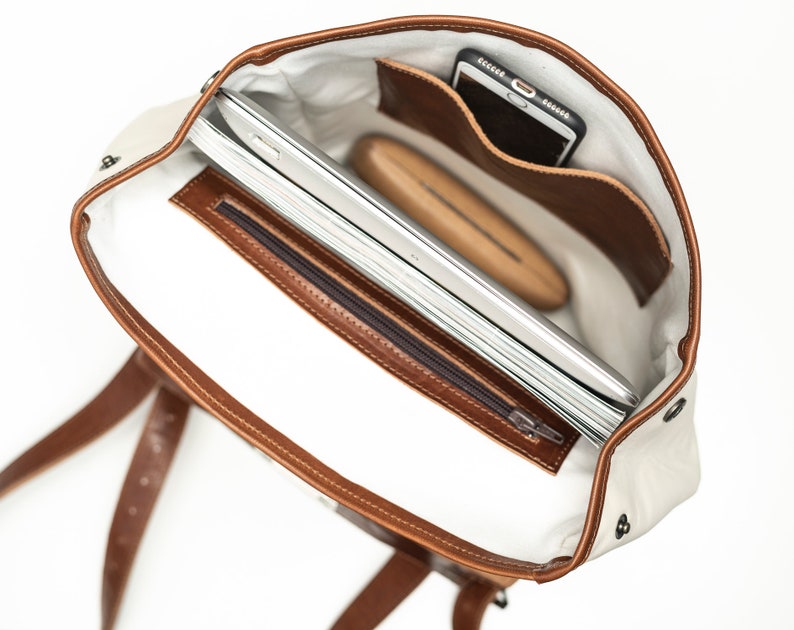 May include: The inside of a brown leather and white canvas backpack. The backpack has a zippered pocket and a large compartment that can fit a laptop and other items. The backpack also has a small pocket for a phone.