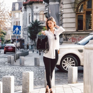 May include: A woman wearing a white button-down shirt, black pants, and brown shoes is walking down a city street. She is carrying a black backpack with a zippered pocket.