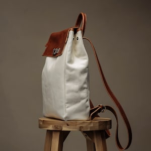 May include: A white canvas backpack with brown leather accents and a buckle closure. The backpack is sitting on a wooden stool.