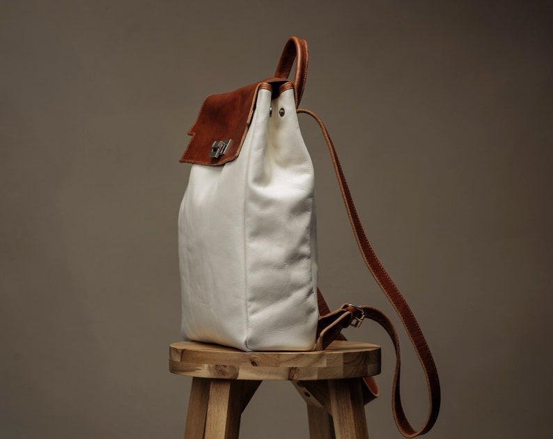 May include: A white canvas backpack with brown leather accents and a buckle closure. The backpack is sitting on a wooden stool.
