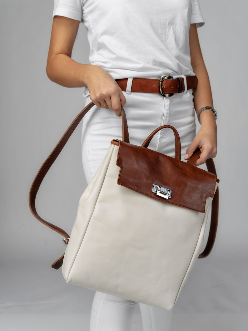 May include: A white leather backpack with a brown leather flap and a silver buckle. The backpack has a brown leather strap.