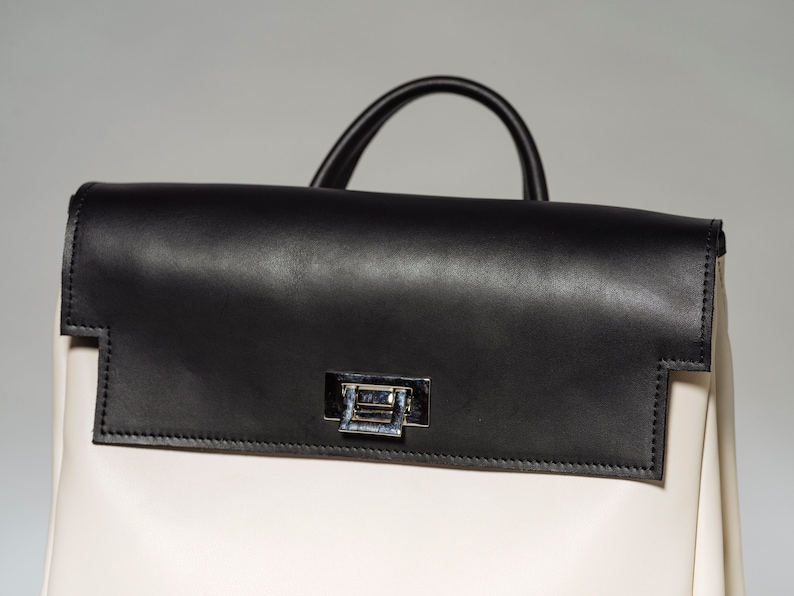 May include: A white leather backpack with a black leather flap and a silver clasp. The backpack has a top handle and a zipper closure.