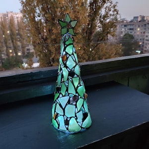 Pode incluir: Uma &aacute;rvore de Natal decorativa feita de pe&ccedil;as de vidro verde e transparente. A &aacute;rvore &eacute; encimada por uma estrela verde e fica sobre uma superf&iacute;cie escura. A &aacute;rvore &eacute; iluminada, criando um efeito brilhante. O fundo mostra uma paisagem urbana.