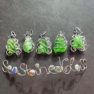 May include: Five green sea glass Christmas tree ornaments wrapped in silver wire. The word "Seaglass" is spelled out in silver wire below the ornaments.