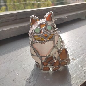 May include: A stained glass cat figurine with brown, white, and green glass pieces. The cat is sitting with its head tilted slightly to the side.