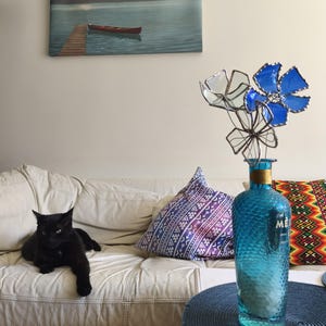 May include: A black cat lounges on a white leather sofa, with patterned cushions. A blue glass bottle with a textured surface holds metal flowers. A painting of a lake scene hangs on the wall.