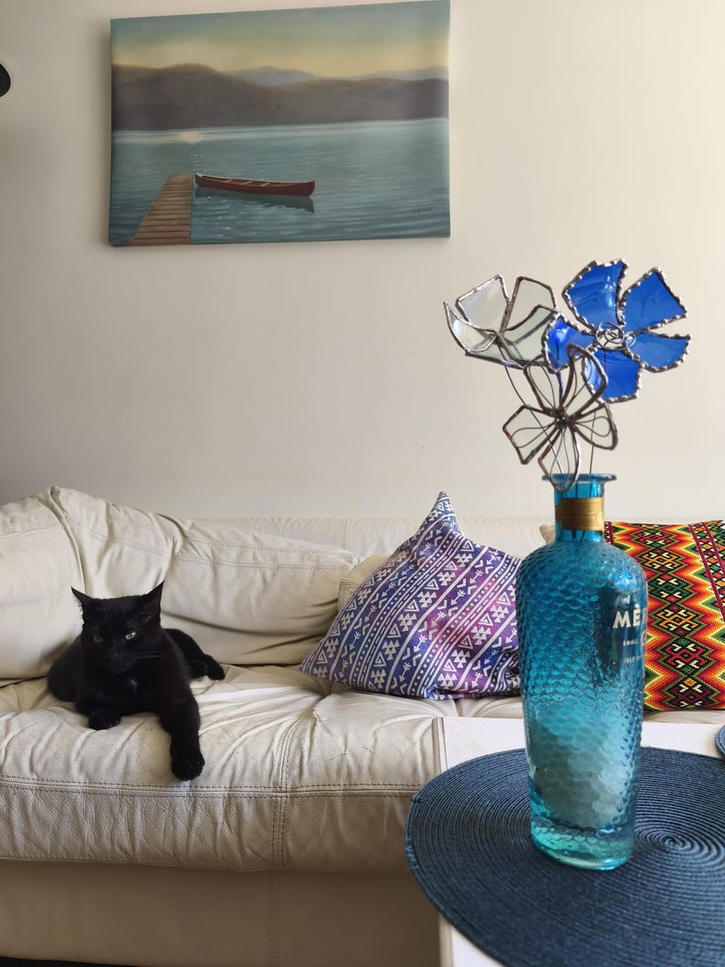 May include: A black cat lounges on a white leather sofa, with patterned cushions. A blue glass bottle with a textured surface holds metal flowers. A painting of a lake scene hangs on the wall.