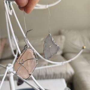 May include: A handcrafted mobile featuring sea glass pieces in shades of pink, blue, and clear, encased in silver wire. The mobile is suspended from a white metal frame, creating a delicate and airy aesthetic. The sea glass pieces vary in size and shape.