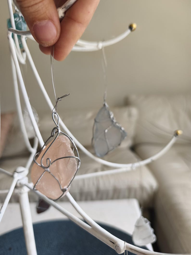 May include: A handcrafted mobile featuring sea glass pieces in shades of pink, blue, and clear, encased in silver wire. The mobile is suspended from a white metal frame, creating a delicate and airy aesthetic. The sea glass pieces vary in size and shape.