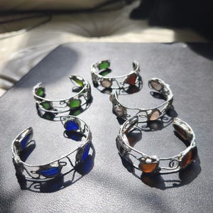 May include: Five silver-toned cuff bracelets with colorful glass accents. The bracelets feature green, blue, orange, and pink glass pieces. The open-ended design allows for easy wear. The bracelets are displayed on a dark surface.