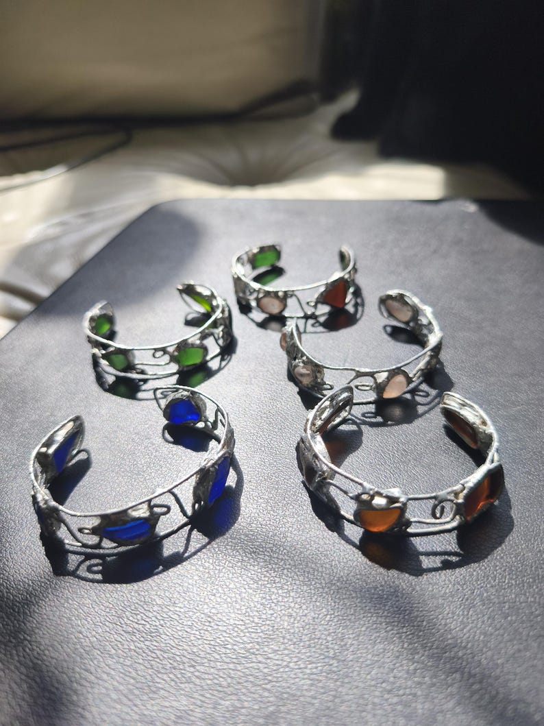 May include: Five silver-toned cuff bracelets with colorful glass accents. The bracelets feature green, blue, orange, and pink glass pieces. The open-ended design allows for easy wear. The bracelets are displayed on a dark surface.