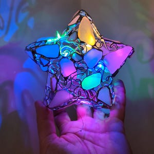 Könnte beinhalten: Ein sternförmiger Glasanhänger mit einem Drahtrahmen und bunten Glasstücken im Inneren. Der Anhänger ist mit mehrfarbigen LED-Lichtern beleuchtet, die einen farbenfrohen und festlichen Glanz erzeugen.