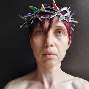 May include: A crown-shaped headpiece constructed from silver-toned metal and small, angular pieces of stained glass in vibrant shades of red, blue, green, and yellow. The structure features sharp, protruding points arranged in a circular band.
