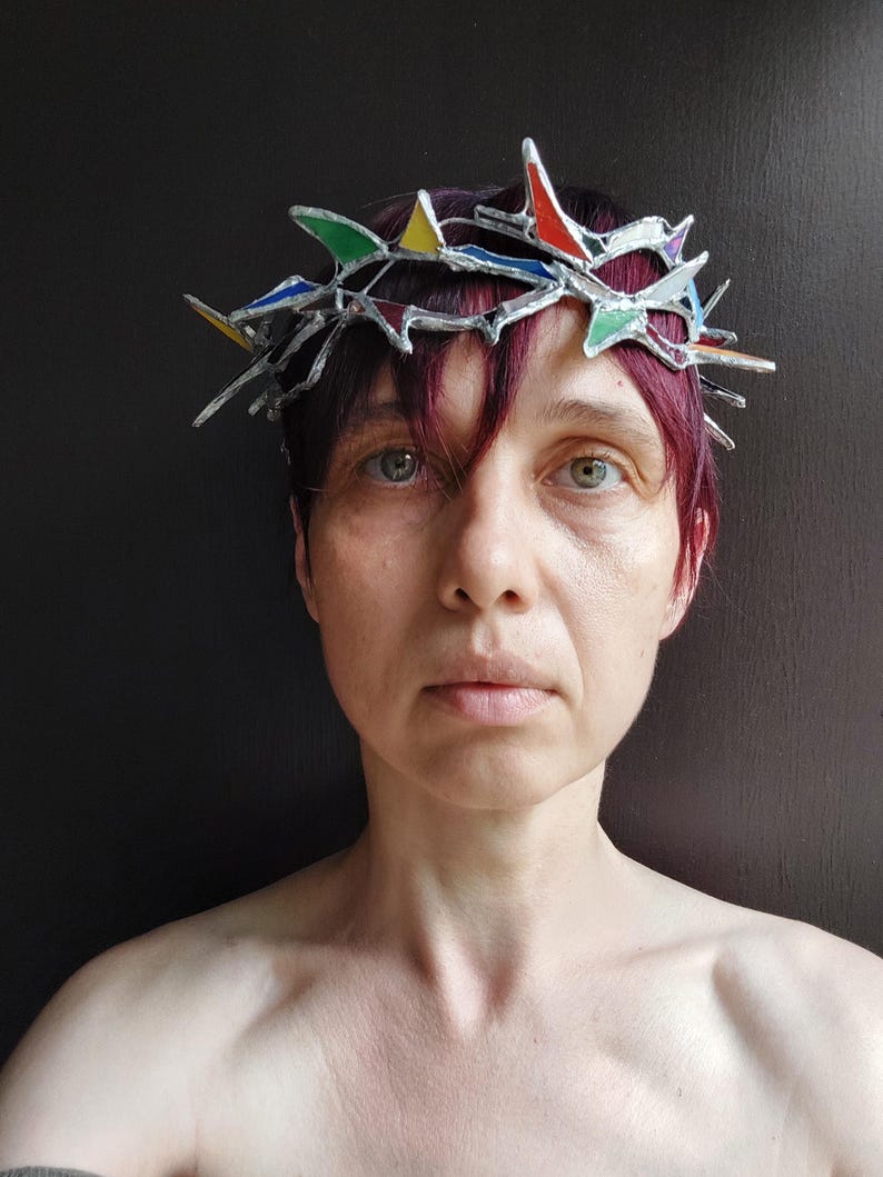 May include: A crown-shaped headpiece constructed from silver-toned metal and small, angular pieces of stained glass in vibrant shades of red, blue, green, and yellow. The structure features sharp, protruding points arranged in a circular band.