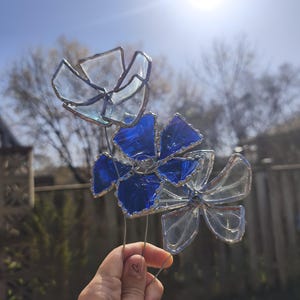 May include: Three stained glass flowers with clear and blue petals, set against a bright sky. The flowers are made with a silver-coloured metal frame and wire stems, creating a delicate garden ornament.