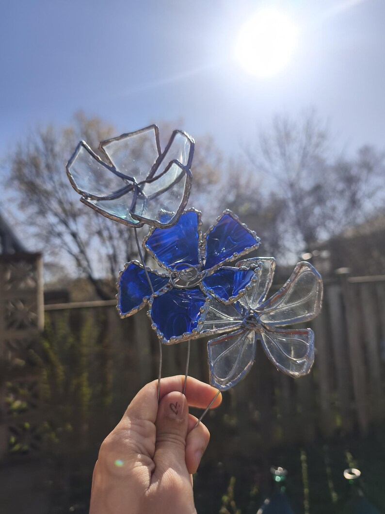 May include: Three stained glass flowers with clear and blue petals, set against a bright sky. The flowers are made with a silver-coloured metal frame and wire stems, creating a delicate garden ornament.