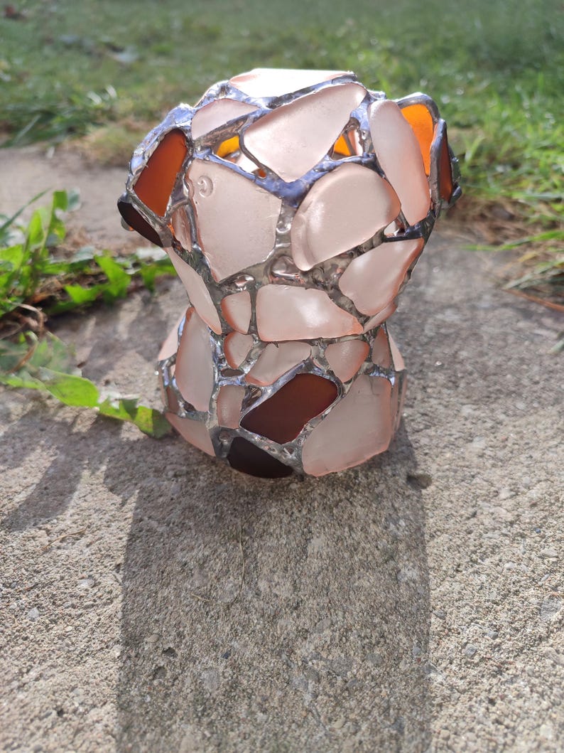 May include: A decorative vase made of sea glass, with a pink and brown color scheme. The vase is made of many small pieces of sea glass that have been fused together.