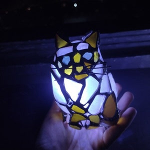 May include: A stained glass cat figurine with yellow, white, and blue glass pieces. The cat is illuminated from within, casting a soft glow.