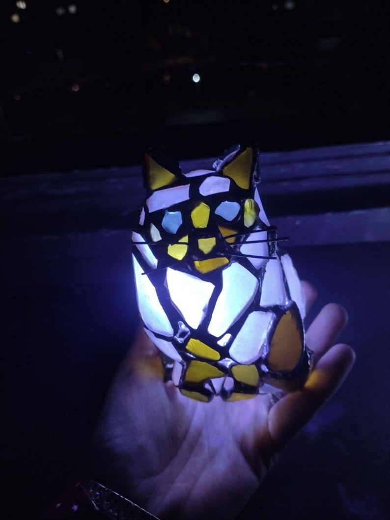 May include: A stained glass cat figurine with yellow, white, and blue glass pieces. The cat is illuminated from within, casting a soft glow.