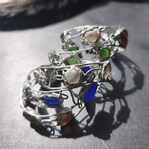 May include: Three silver-toned wire rings with colorful glass stones. The rings feature a delicate, openwork design and are adorned with various colored stones, including pink, green, blue, and orange. The rings are stacked on top of each other.