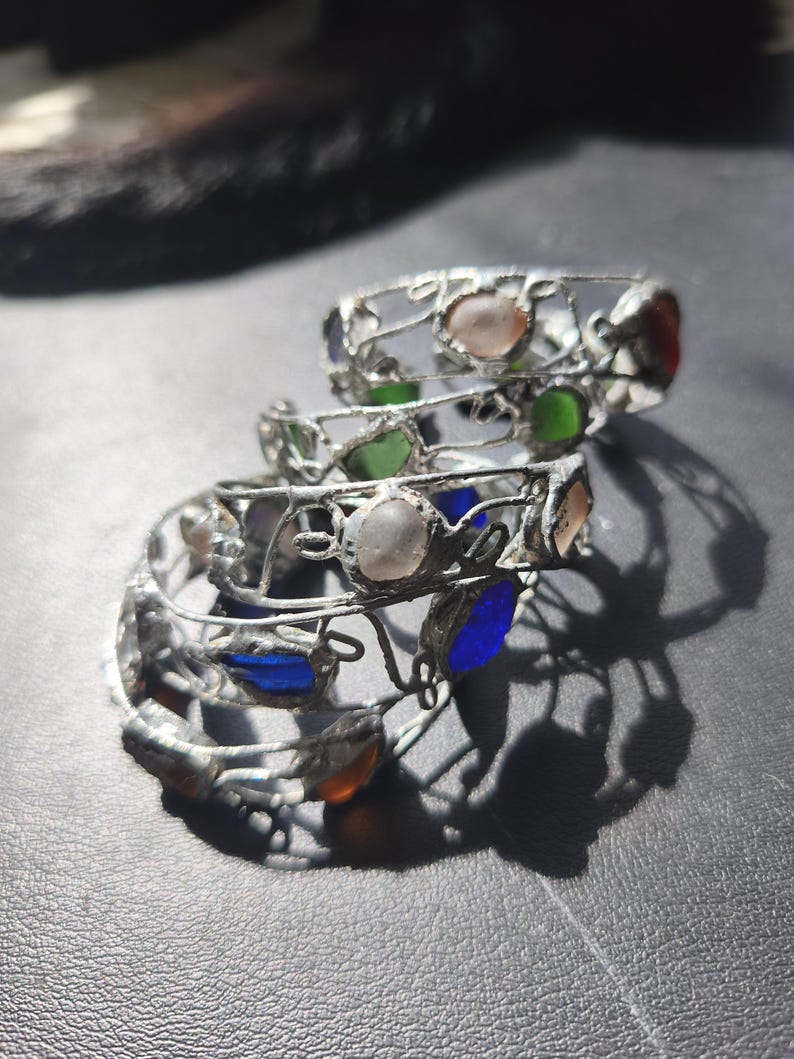 May include: Three silver-toned wire rings with colorful glass stones. The rings feature a delicate, openwork design and are adorned with various colored stones, including pink, green, blue, and orange. The rings are stacked on top of each other.