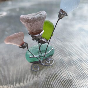 May include: A handcrafted sculpture of sea glass flowers. The sculpture has a green base with a swirling wire design. The flowers are made of various colours of sea glass, including pink, green, and light blue. The stems are thin metal wires.