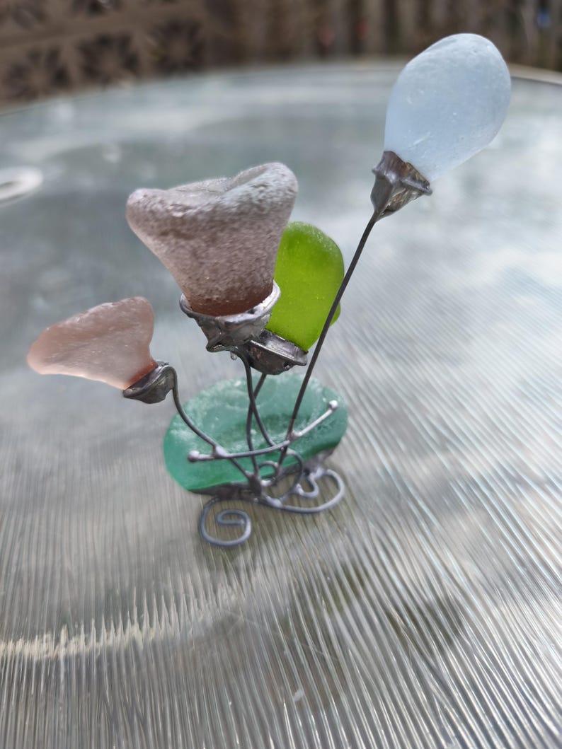 May include: A handcrafted sculpture of sea glass flowers. The sculpture has a green base with a swirling wire design. The flowers are made of various colours of sea glass, including pink, green, and light blue. The stems are thin metal wires.
