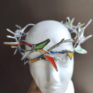 May include: A crown-shaped headpiece constructed from silver-toned metal branches and small, triangular stained glass inserts in vibrant shades of red, green, blue, and yellow, arranged in a jagged, thorn-like pattern around the circular frame.