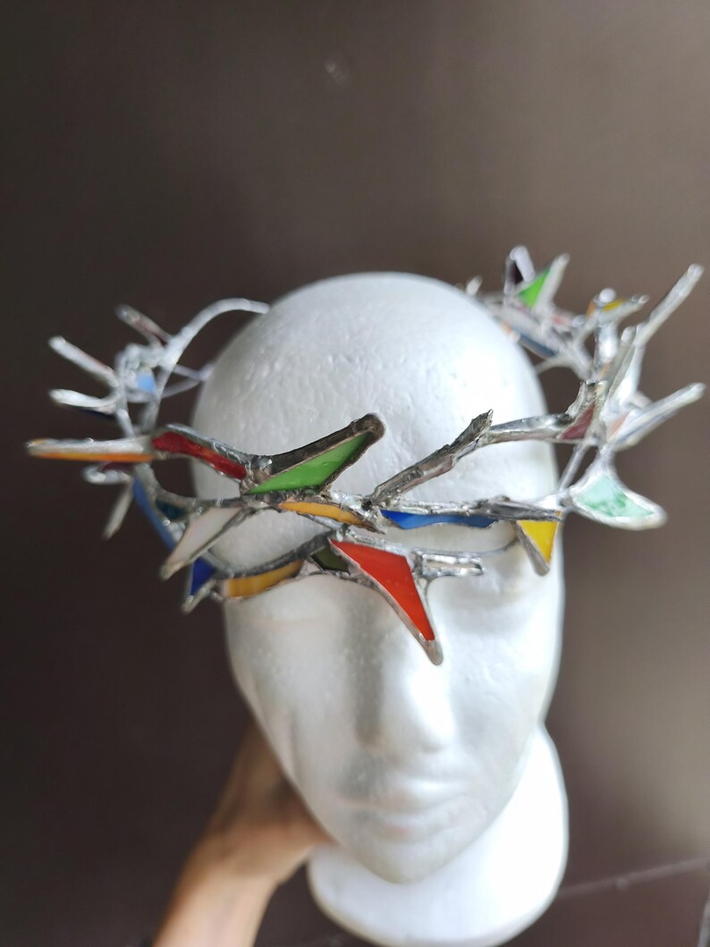 May include: A crown-shaped headpiece constructed from silver-toned metal branches and small, triangular stained glass inserts in vibrant shades of red, green, blue, and yellow, arranged in a jagged, thorn-like pattern around the circular frame.