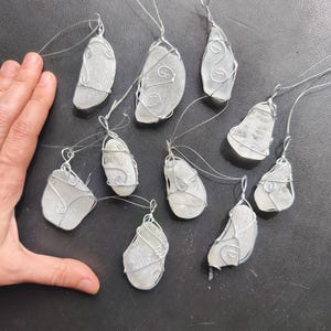 May include: A collection of ten sea glass pendants, each with a unique shape and wrapped in silver-colored wire. The pendants are suspended from thin, clear cords, suggesting they are designed to be hung or worn. The sea glass has a frosted, opaque appearance.