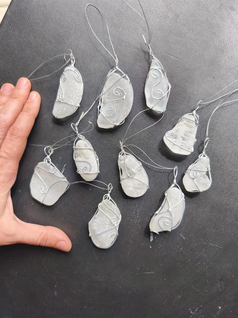 May include: A collection of ten sea glass pendants, each with a unique shape and wrapped in silver-colored wire. The pendants are suspended from thin, clear cords, suggesting they are designed to be hung or worn. The sea glass has a frosted, opaque appearance.