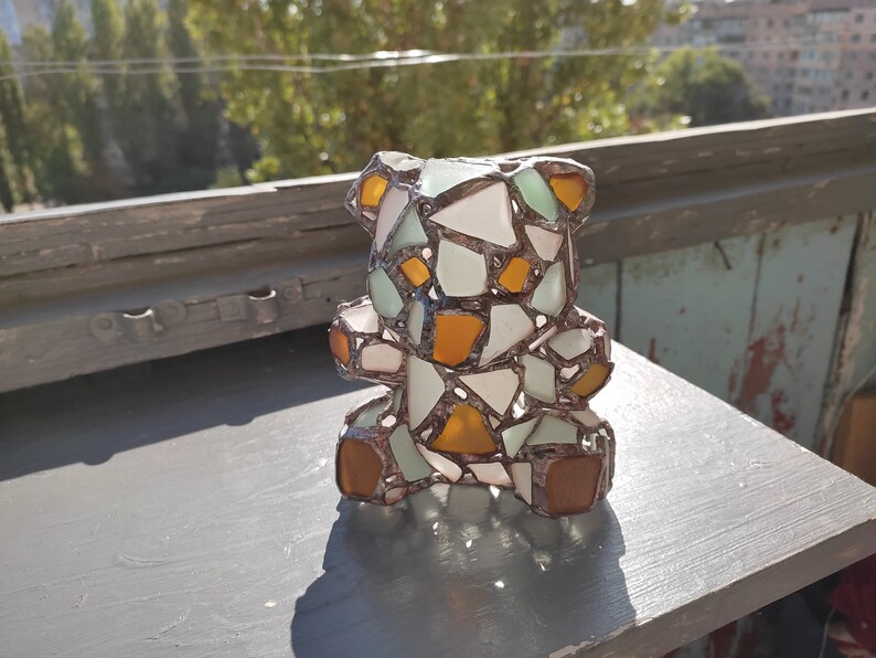 May include: A mosaic teddy bear sculpture composed of various colored glass pieces. The bear features white, orange, and green glass fragments, with brown glass for the feet and ears. The artwork is displayed on a gray surface.