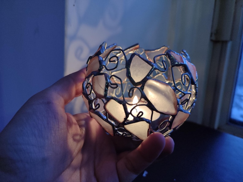May include: A candle holder made of clear glass and silver wire, with a candle lit inside. The holder is shaped like a sphere and has a delicate, intricate design.