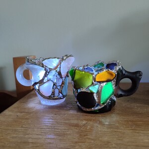 May include: Two small ceramic mugs decorated with colorful mosaic glass pieces. The mugs have a handle and are made in a light blue and black color scheme.