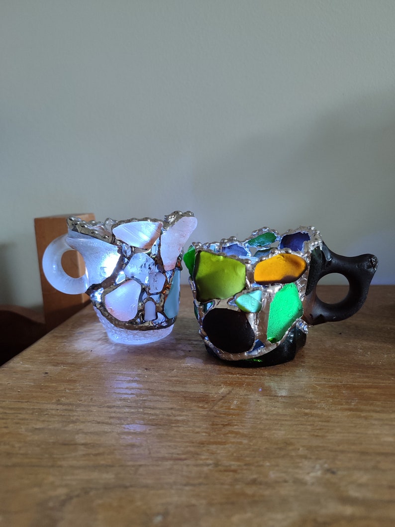 May include: Two small ceramic mugs decorated with colorful mosaic glass pieces. The mugs have a handle and are made in a light blue and black color scheme.