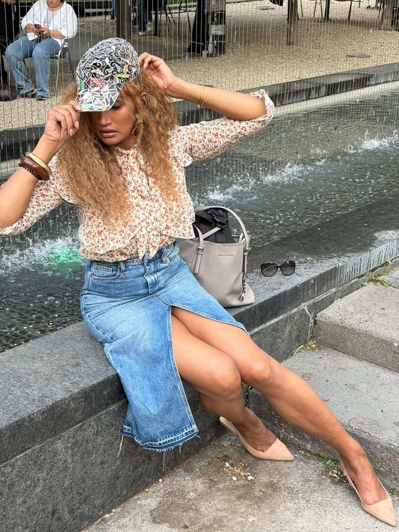 May include: A woman wearing a floral print blouse, a denim skirt with a front slit, and a patterned baseball cap. She is also wearing beige heels and a gray handbag. The background includes a water feature and outdoor seating.