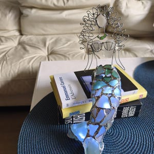 Puede incluir: Una escultura decorativa de una figura de sirena hecha de alambre y vidrio marino. La parte superior del cuerpo est&aacute; formada por alambre plateado con cabeza y pecho de vidrio marino. La cola se compone de fragmentos de vidrio azul y verde.