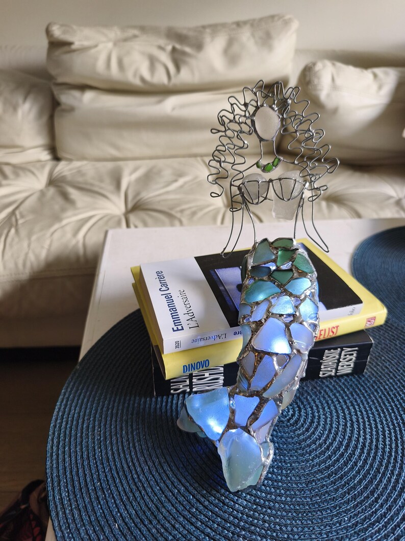 Puede incluir: Una escultura decorativa de una figura de sirena hecha de alambre y vidrio marino. La parte superior del cuerpo est&aacute; formada por alambre plateado con cabeza y pecho de vidrio marino. La cola se compone de fragmentos de vidrio azul y verde.