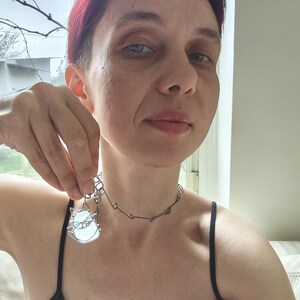 May include: A silver-toned necklace with a pendant featuring a cat design. The pendant has a light blue, oval-shaped stone. The necklace is worn with a black tank top.