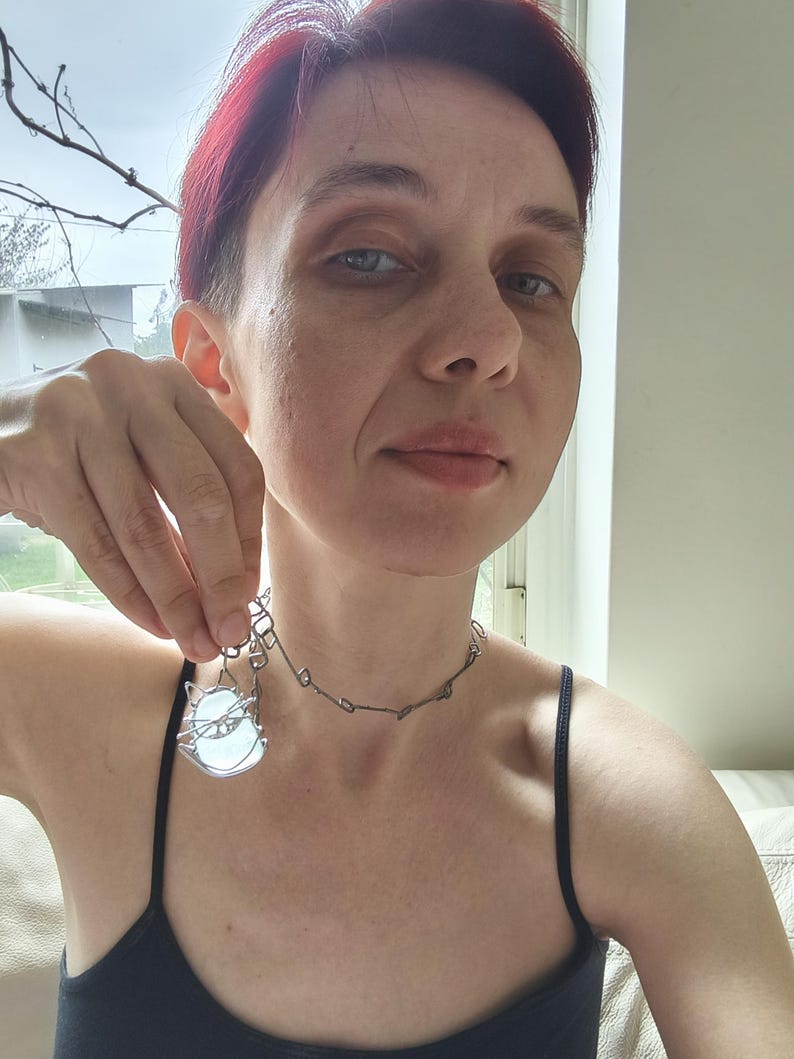 May include: A silver-toned necklace with a pendant featuring a cat design. The pendant has a light blue, oval-shaped stone. The necklace is worn with a black tank top.