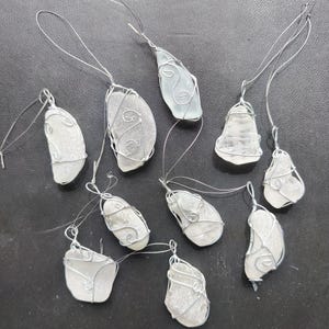 May include: A collection of ten sea glass pendants. Each piece of sea glass is wrapped in silver-colored wire with a loop for hanging. The sea glass varies in shape and color, ranging from clear to frosted white.