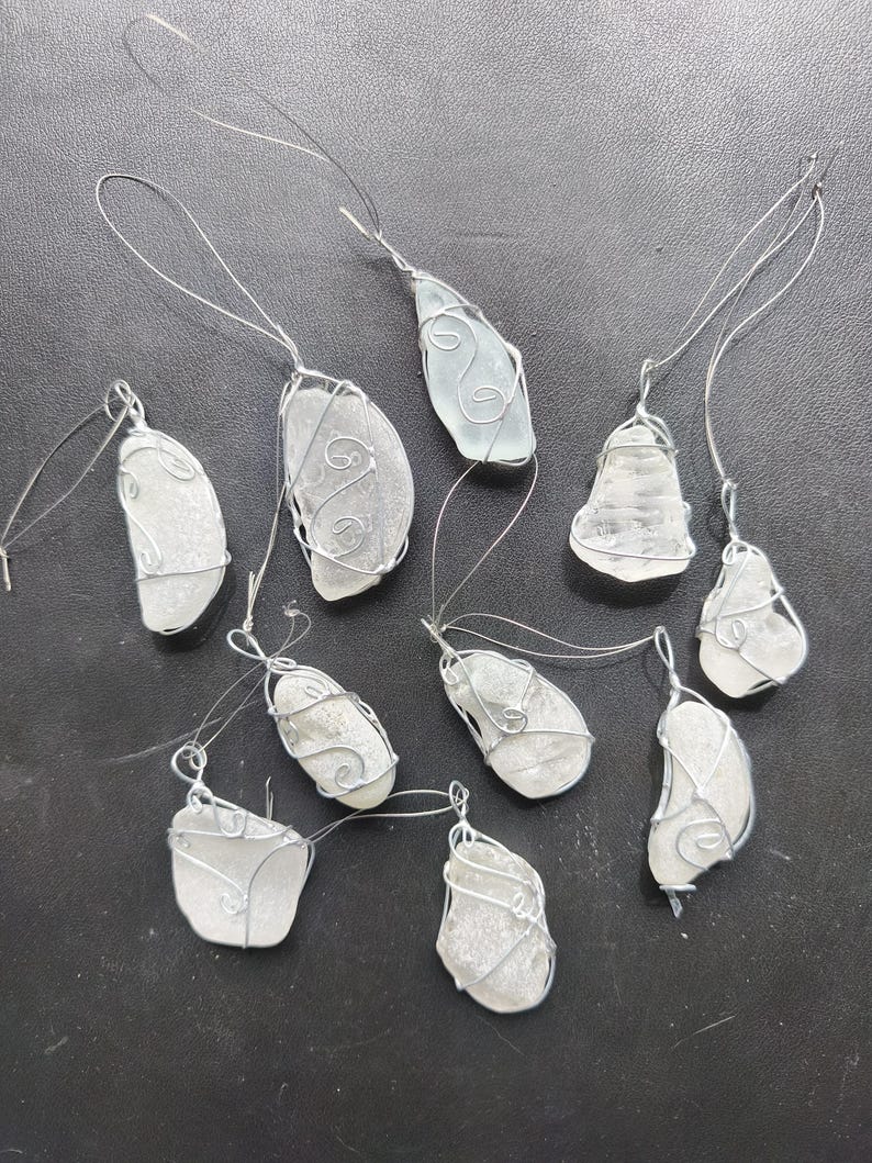 May include: A collection of ten sea glass pendants. Each piece of sea glass is wrapped in silver-colored wire with a loop for hanging. The sea glass varies in shape and color, ranging from clear to frosted white.
