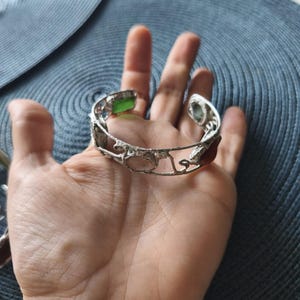 May include: Silver-toned cuff bracelet with an openwork design, featuring various colored gemstones. The bracelet includes green, pink, and brown stones. The bracelet is displayed on a hand, showcasing its intricate details and craftsmanship.