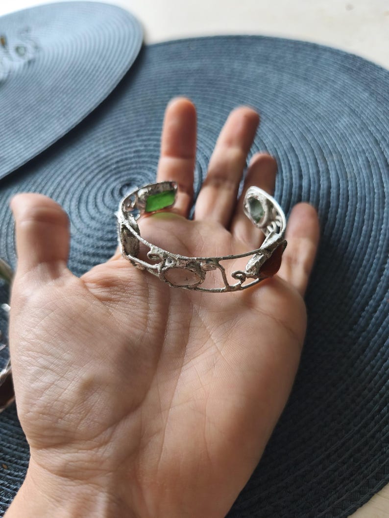 May include: Silver-toned cuff bracelet with an openwork design, featuring various colored gemstones. The bracelet includes green, pink, and brown stones. The bracelet is displayed on a hand, showcasing its intricate details and craftsmanship.