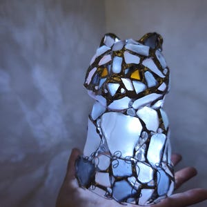 May include: A mosaic cat ornament with an internal light source. The cat is made from irregular pieces of white, blue, and gold-toned glass, with wire detailing. The eyes are a warm yellow colour. The ornament is held in a hand.