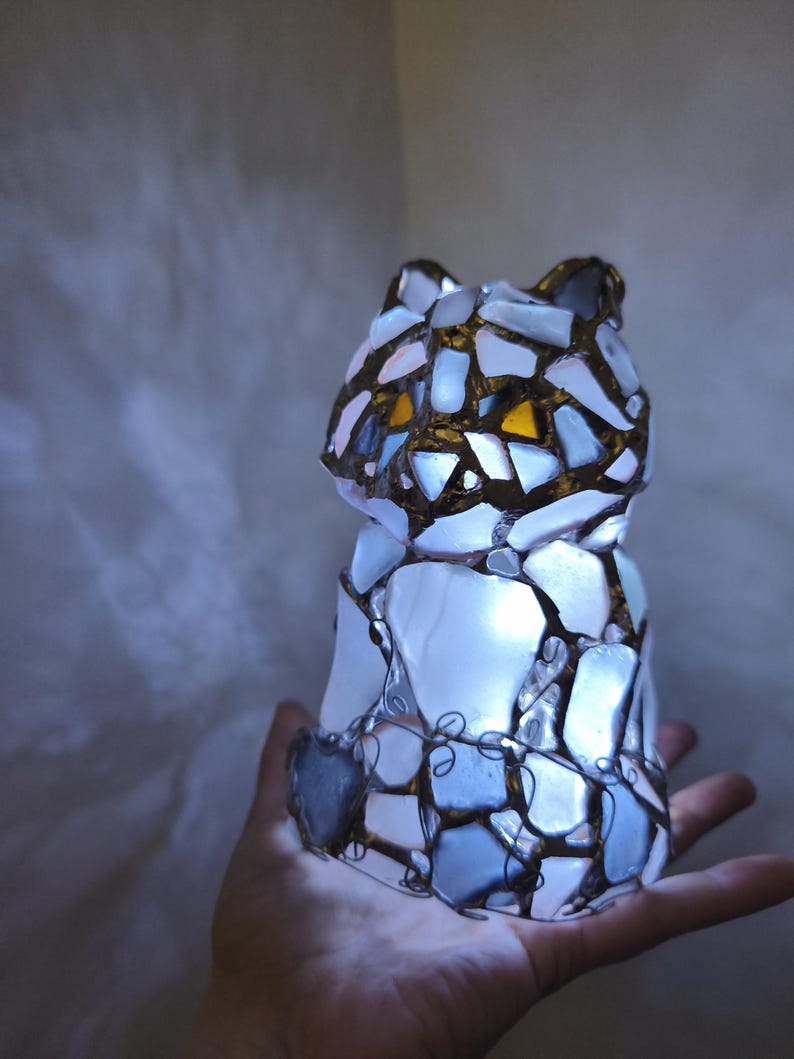 May include: A mosaic cat ornament with an internal light source. The cat is made from irregular pieces of white, blue, and gold-toned glass, with wire detailing. The eyes are a warm yellow colour. The ornament is held in a hand.