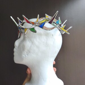 May include: A crown made of stained glass pieces in various colors including red, yellow, blue, green, and purple, joined together with silver-toned metal solder. The crown features sharp, pointed spikes and is displayed on a white mannequin head.